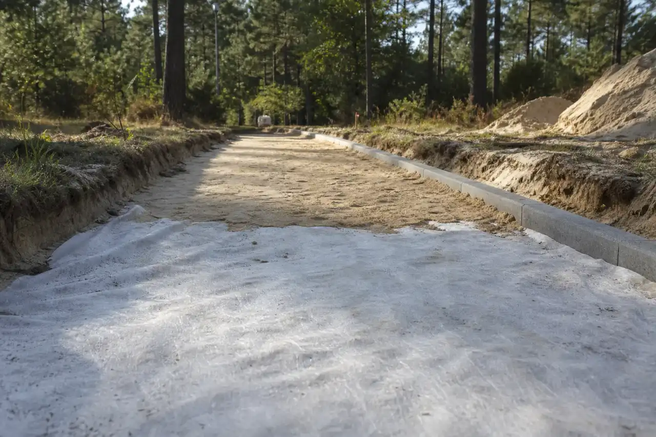 Coupe d'une allée en cours de création : géotextile et fondation compactée visibles sur le sable clair avant la pose des gravillons, bordure alignée.