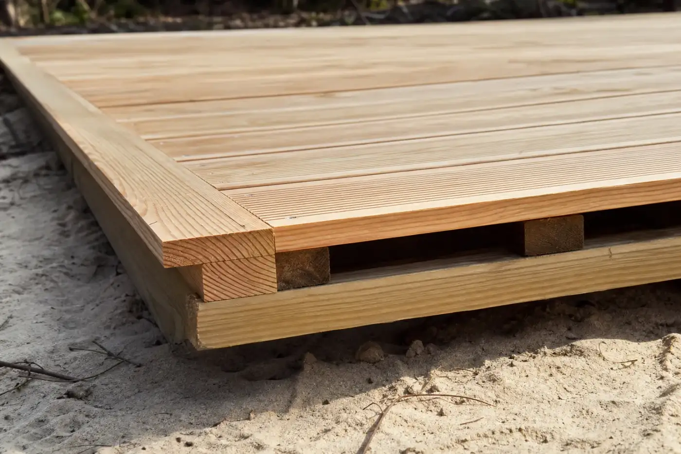 Gros plan sur des lames de terrasse en pin des Landes fraîchement posées, jeu régulier entre lames, structure ventilée visible en rive, sable clair dessous.