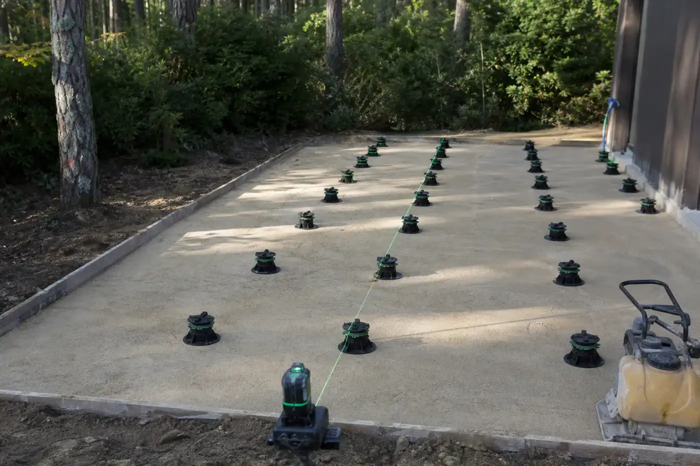 Plateforme de terrasse en cours de préparation sur sol sableux : lit drainant compacté à la plaque vibrante, plots réglables alignés au laser avant pose.